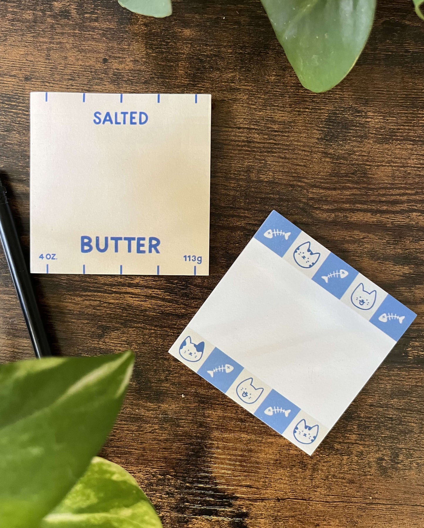 Two square sticky note pads on a wooden surface. The left sticky note mimics butter packaging with "Salted Butter" text. The cat stick notes on the right has blue borders featuring cat faces and fish bones.