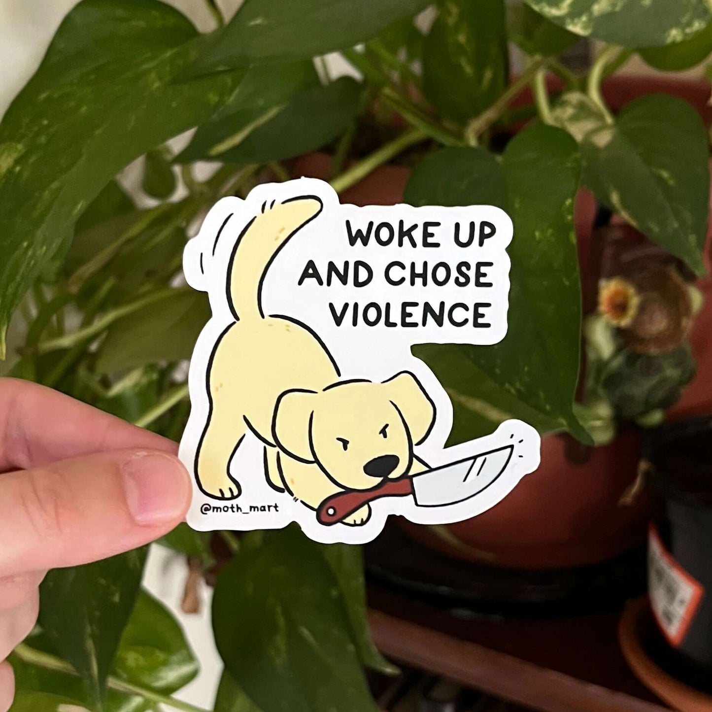 Hand holding a sticker with a dog and text 'Woke up and chose violence' in front of plants and books.