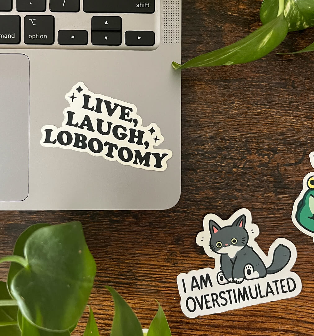 Sticker on a laptop corner that reads "Live, Laugh, Lobotomy" with a second sticker depicting a cat that reads "I am overstimulated".