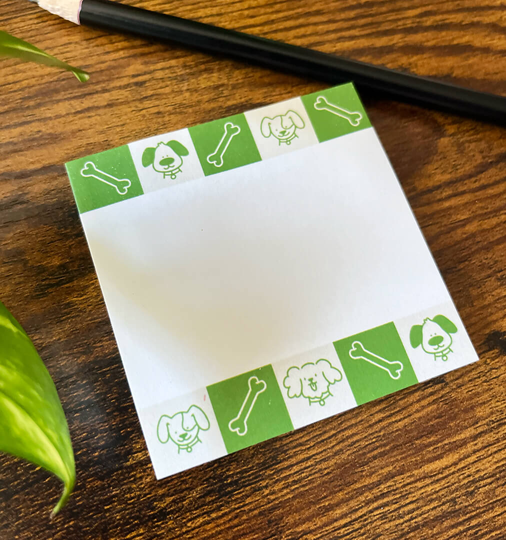 Angled sticky notepad on wood background depicting green and beige squares featuring cartoon dogs and bones. Pencil off to the side.