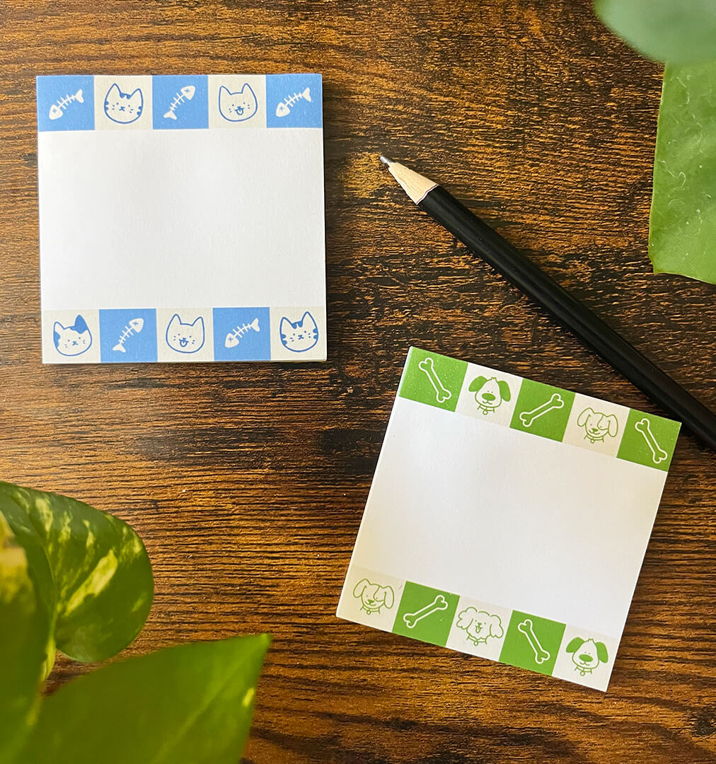 Two decorative sticky notepads on a wooden surface. The top notepad has blue cat and fish bone patterns, while the bottom one has green dog and bone designs. A pencil and plant leaves are nearby.