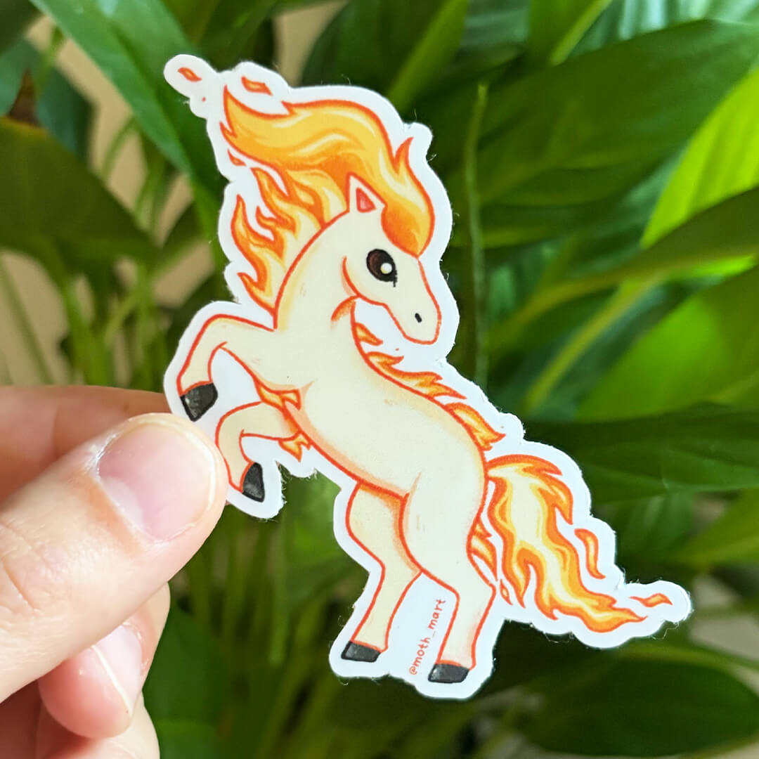 A hand holds a die-cut sticker of a cartoon white horse with orange and yellow flames for a mane and tail, set against a background of green houseplants.
