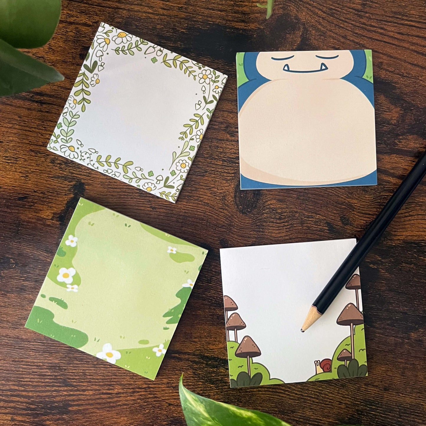 Four decorative sticky notes on a wooden table. Designs include floral, sleeping normal type, green landscape, and mushroom with snail. A pencil is nearby.