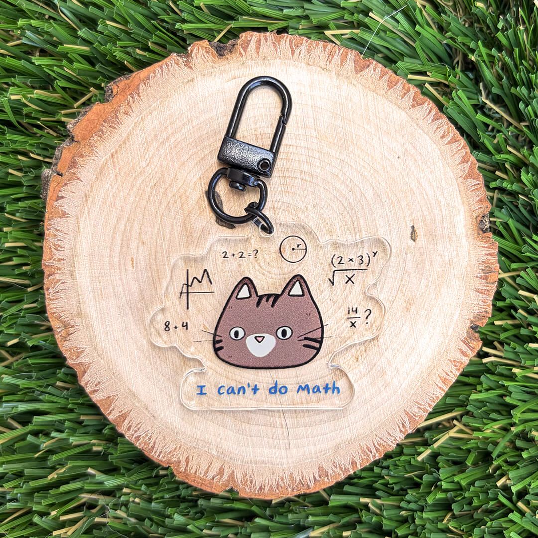 A clear acrylic keychain featuring a cute cartoon cat with math problems and the text 'I can't do Math' in blue, attached to a black metal clasp. The keychain is displayed on a round wood slice over a green grass background.