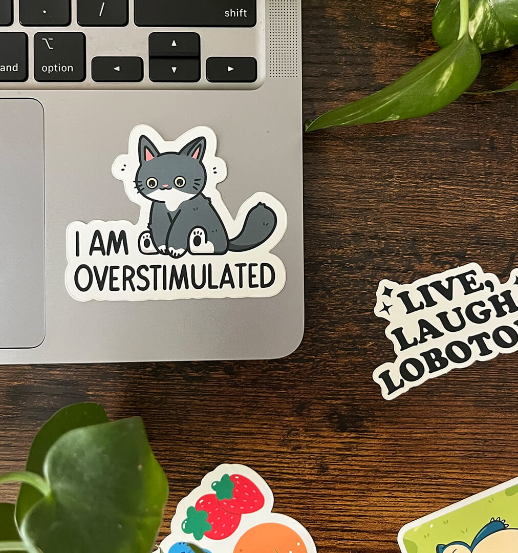 A laptop with an overstimulated cat sticker with a cartoon cat with wide eyes sits above the words "I am overstimulated." Three stickers sit on the border.
