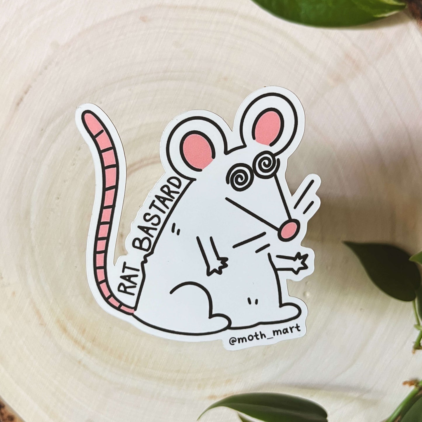Cartoon rat sticker with text that says "rat bastard" on a wooden surface with leaves