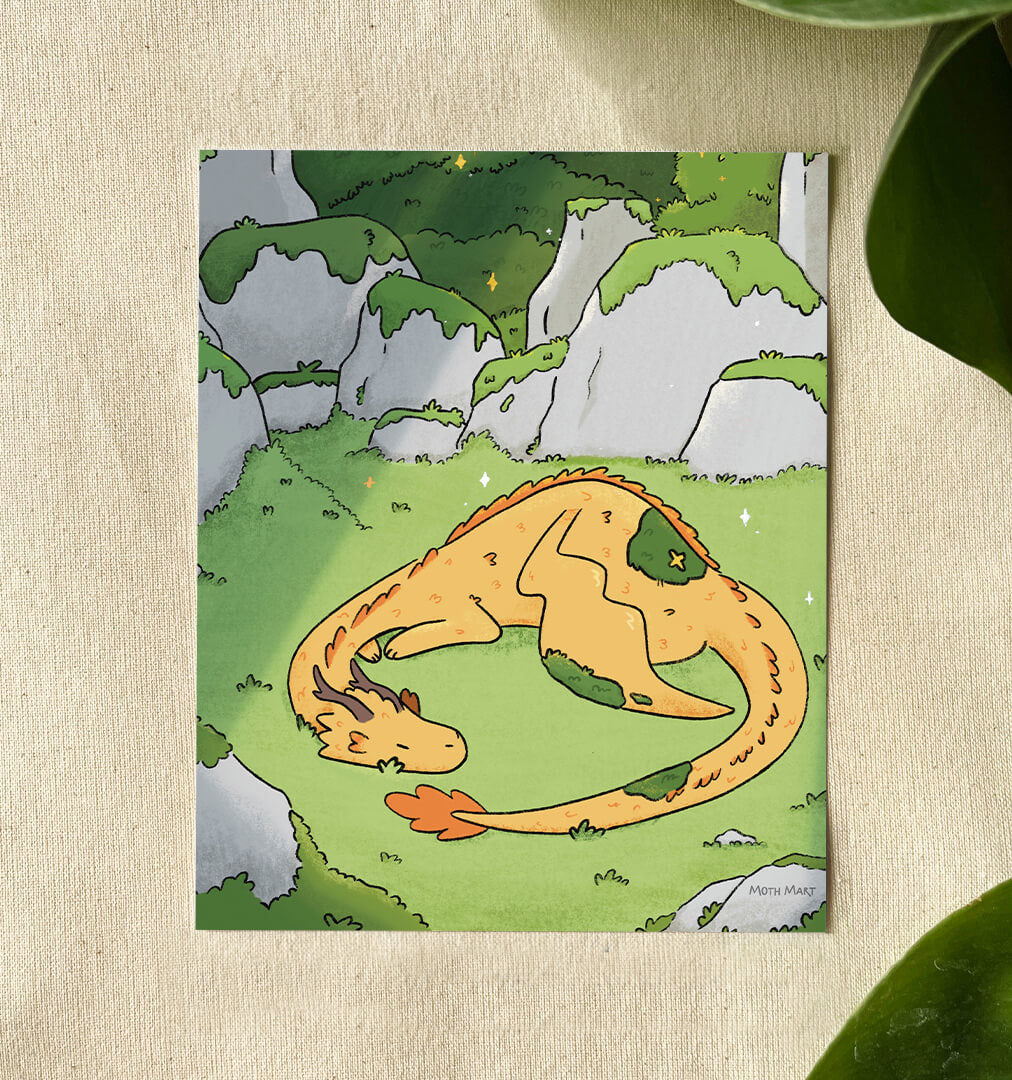 A vertical art print featuring a whimsical illustration of a sleeping orange dragon curled up in a grassy green landscape with gray rocks, displayed on a textured beige fabric background with green leaves in the corners.