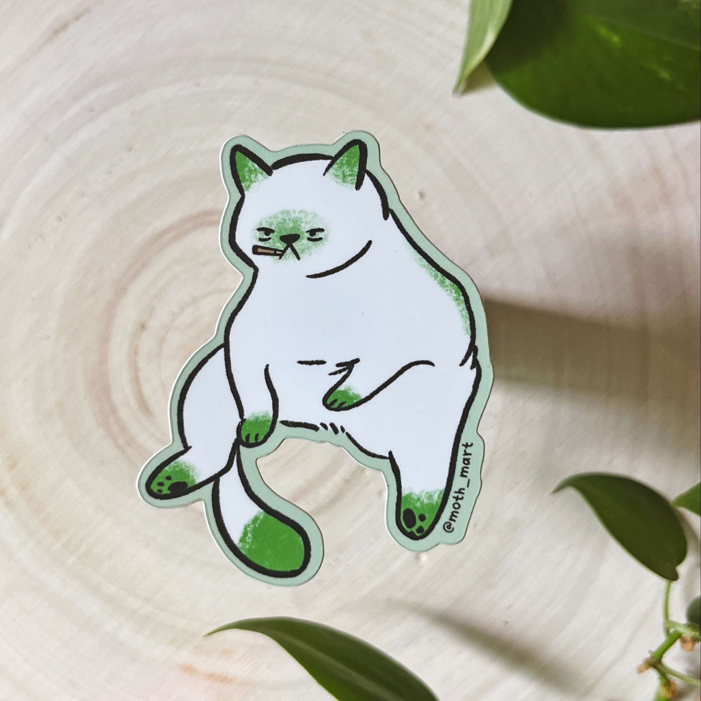 White smoking cat sticker featuring green accents on a wooden surface with leaves.