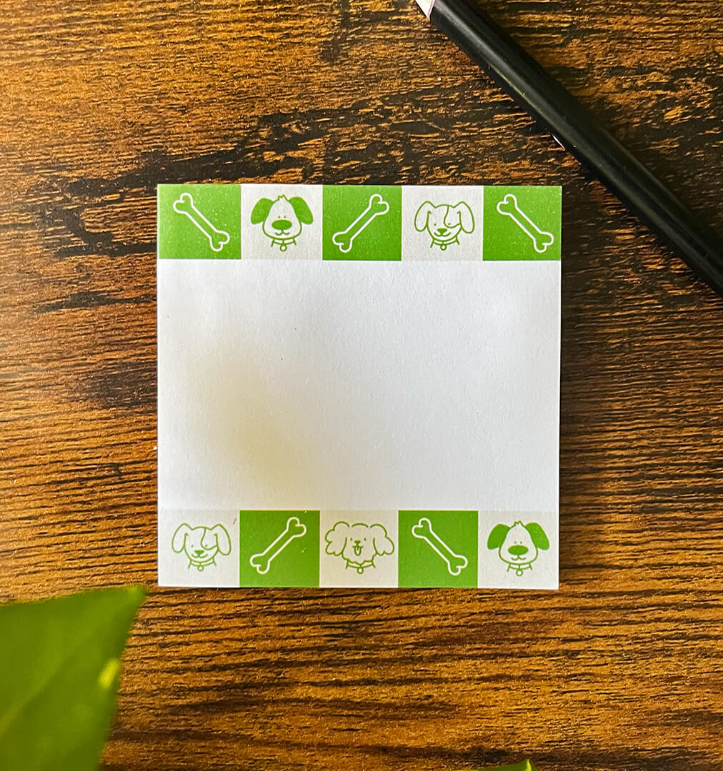 Sticky notepad on wood background depicting green and beige squares featuring cartoon dogs and bones. Pencil off to the side.