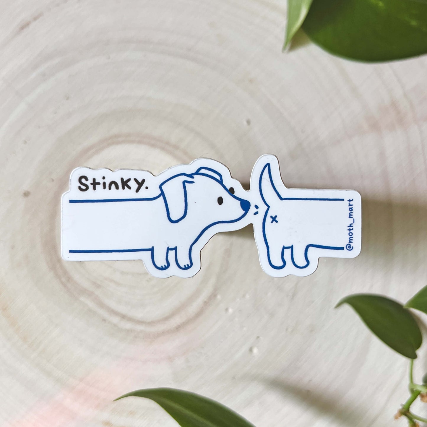 Sticker of a dog sniffing another dog's butt with 'Stinky' text on a wooden surface with green leaves.