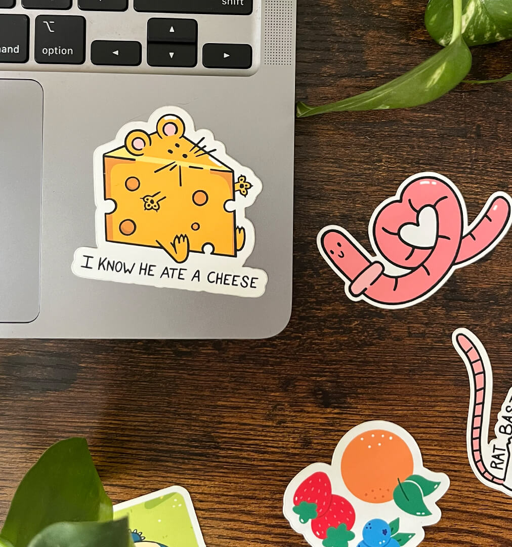 Sticker of a mouse in the shape of a block of yellow cheese with the text "I know he ate a cheese" on the corner of a laptop. Three stickers sit to the side. 