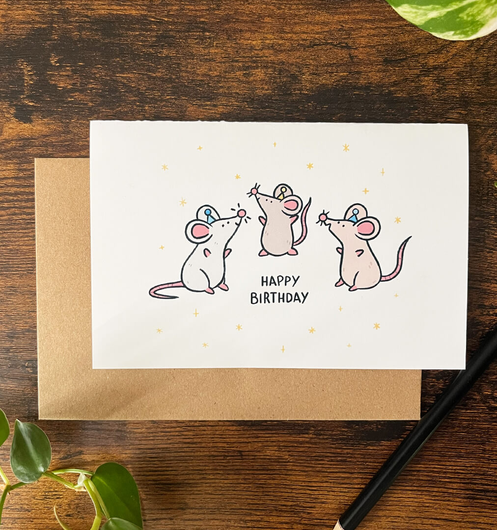 Cartoon on a greeting card of 3 brown and grey rats wearing party hats. They are surrounded by stars with the text "Happy Birthday" on a wood table. 