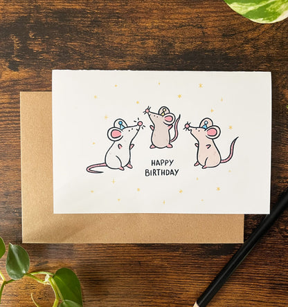Cartoon on a greeting card of 3 brown and grey rats wearing party hats. They are surrounded by stars with the text "Happy Birthday" on a wood table. 