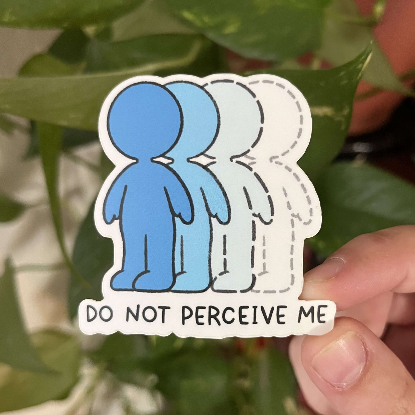 Stickers of blue cartoon figures in gradient colors with text "Do Not Perceive Me," held by hand against foliage. 