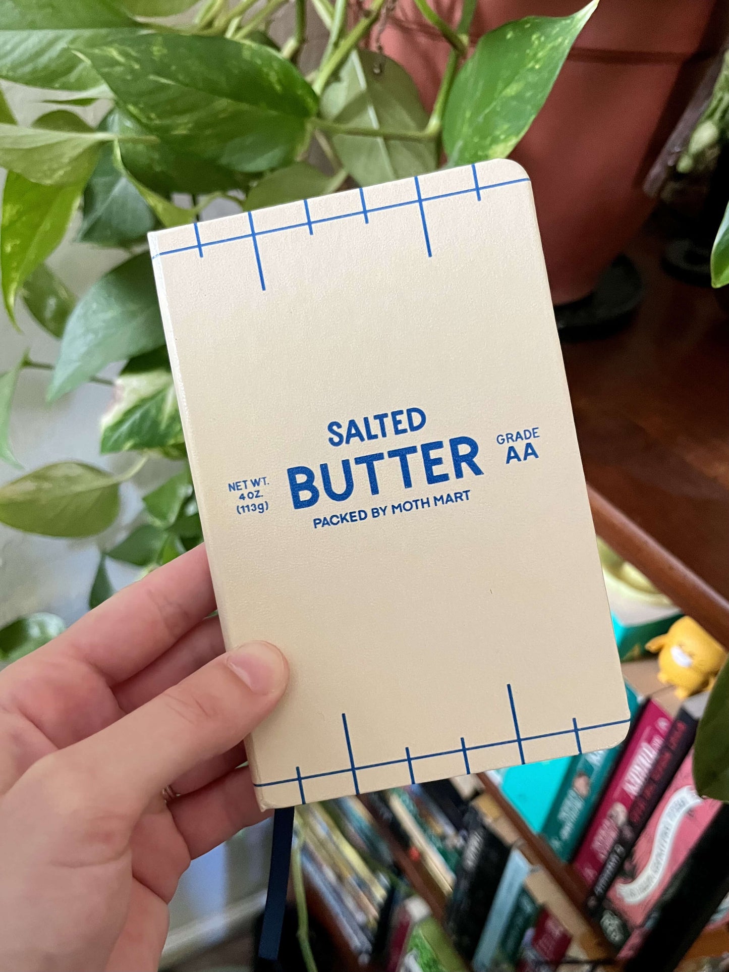 A hand holds a notebook designed to look like a salted butter package, with text and grid lines. In the background, there are green leaves and books.