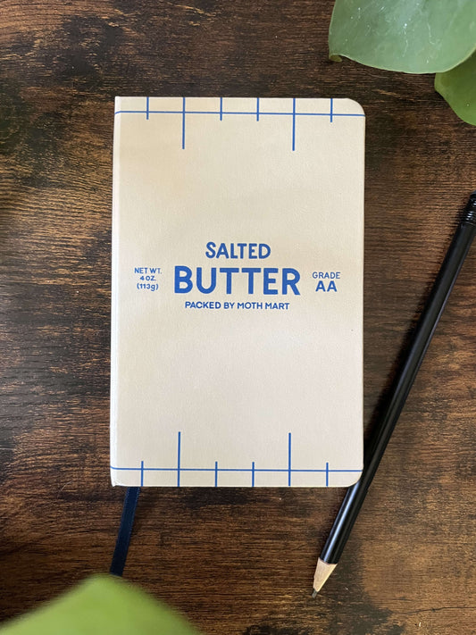 Butter-themed notebook on wooden table. The notebook cover resembles a butter package, featuring blue text and markings that read "Salted Butter". A black pencil lies nearby.