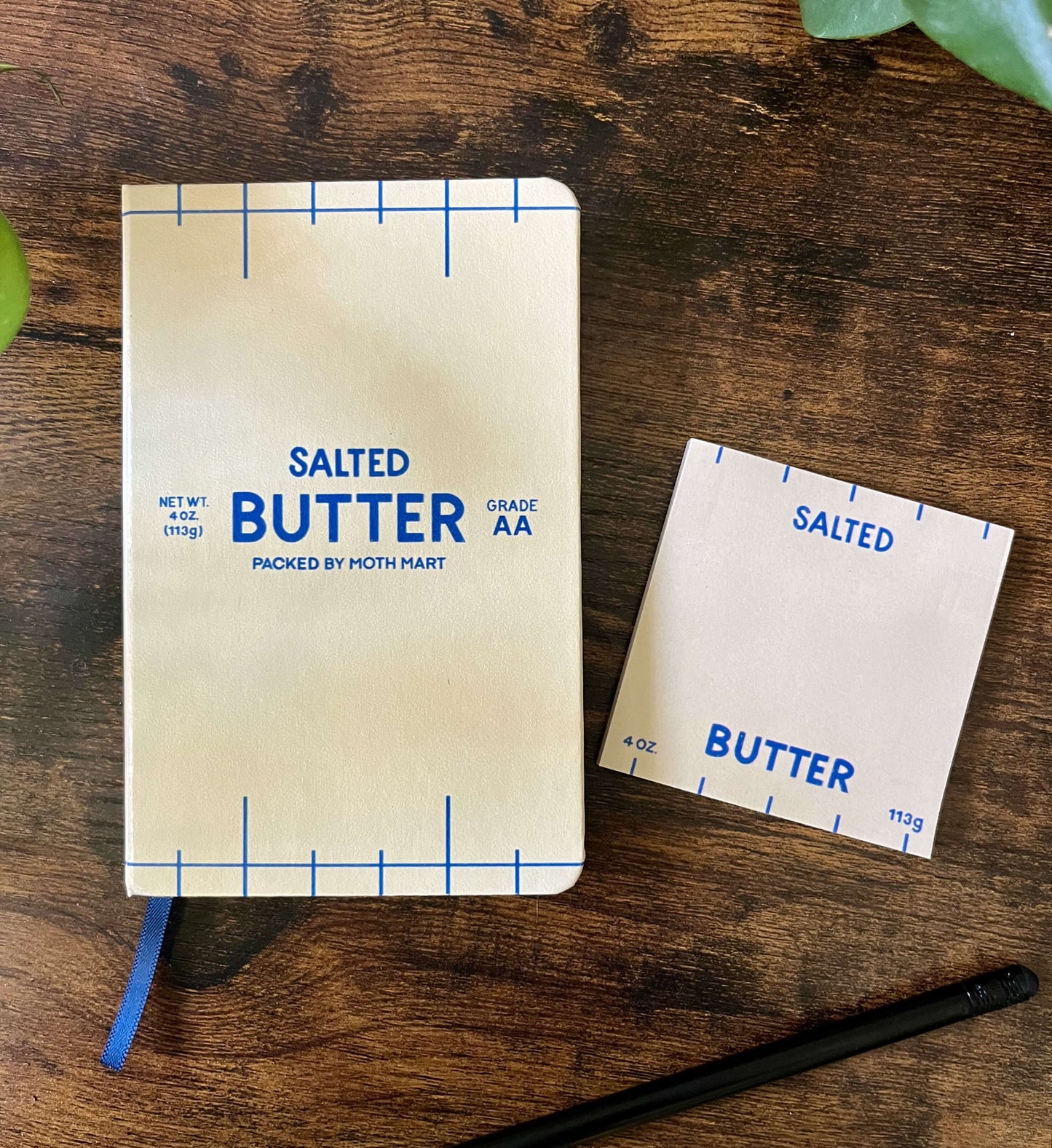 Butter-themed notebook and sticky notes on wooden table. The notebook cover resembles a butter package, featuring blue text and markings that read "Salted Butter". A black pencil lies nearby.