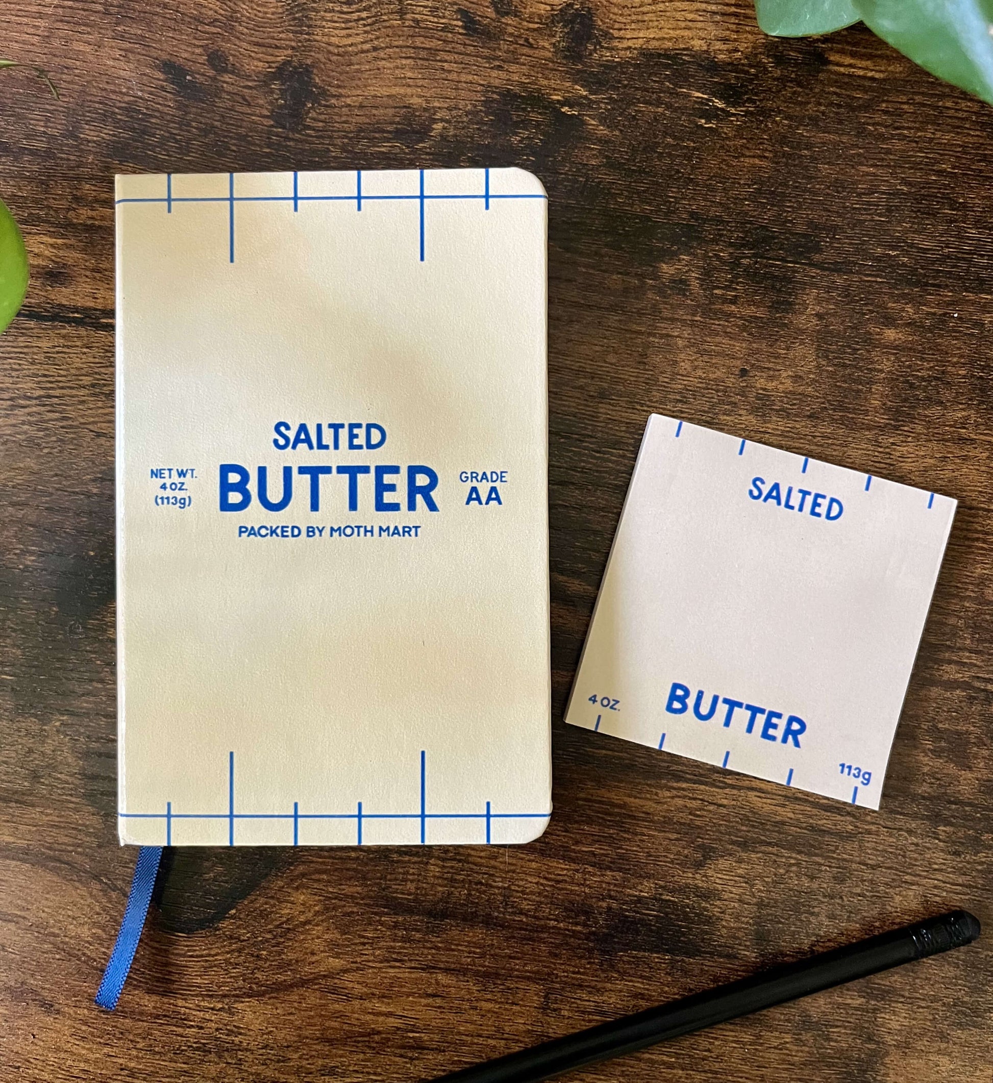 Butter-themed notebook and sticky notes on wooden table. The notebook cover resembles a butter package, featuring blue text and markings that read "Salted Butter". A black pencil lies nearby.