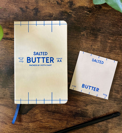 Butter-themed notebook and sticky notes on wooden table. The notebook cover resembles a butter package, featuring blue text and markings that read "Salted Butter". A black pencil lies nearby.