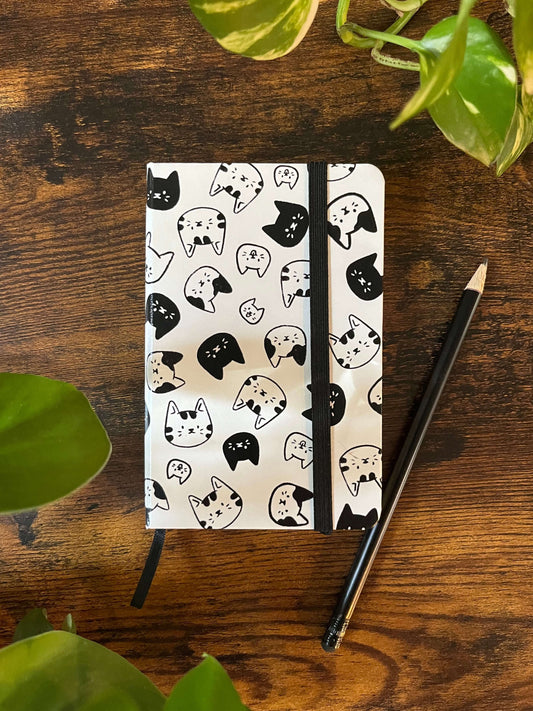A notebook with a white cover featuring a pattern of various black and white cat faces, tied with a black elastic band and sitting on a wood table. 