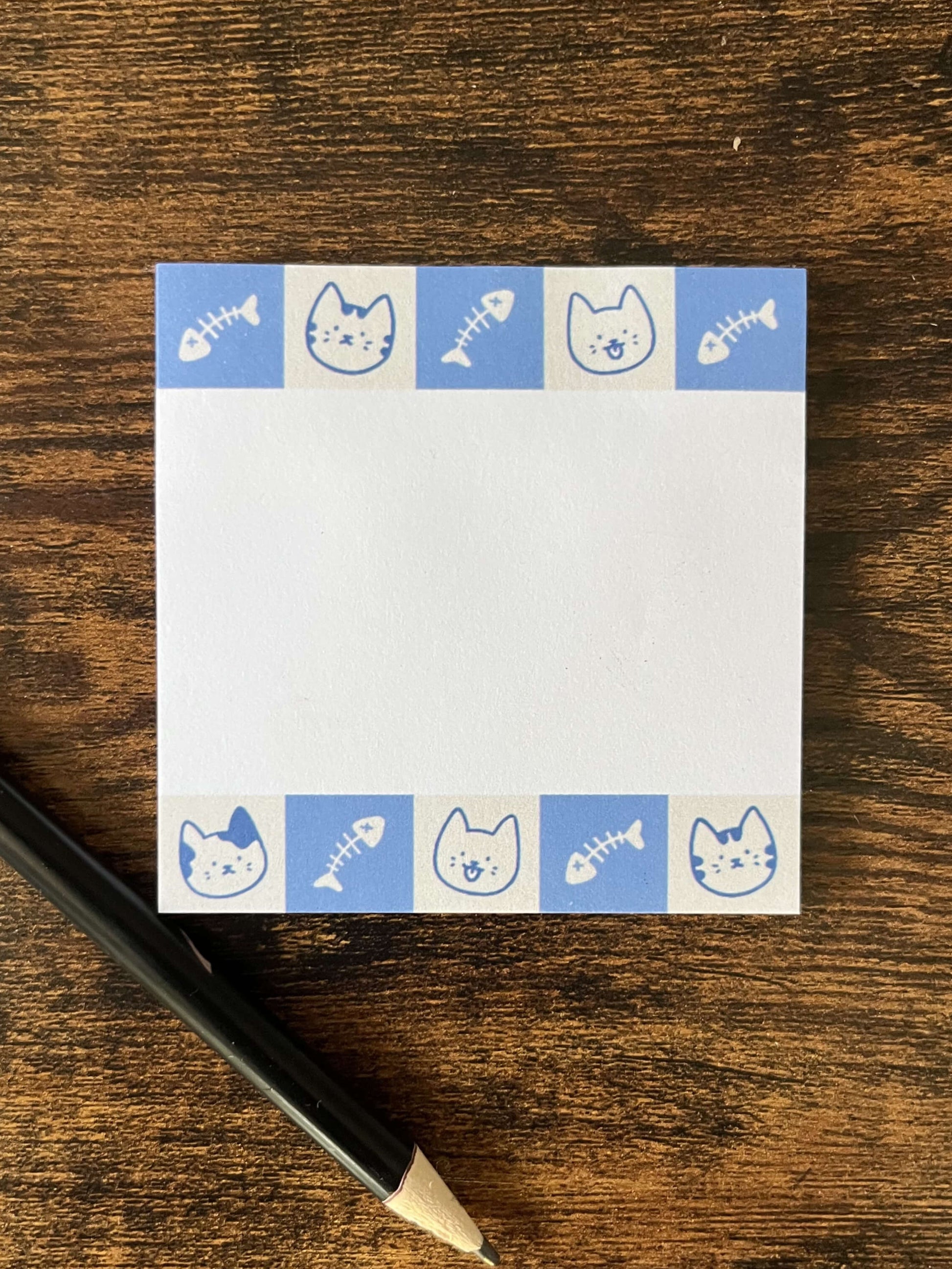 One square sticky notepad on a wood back. The sticky note has blue borders featuring cat faces and fish bones.