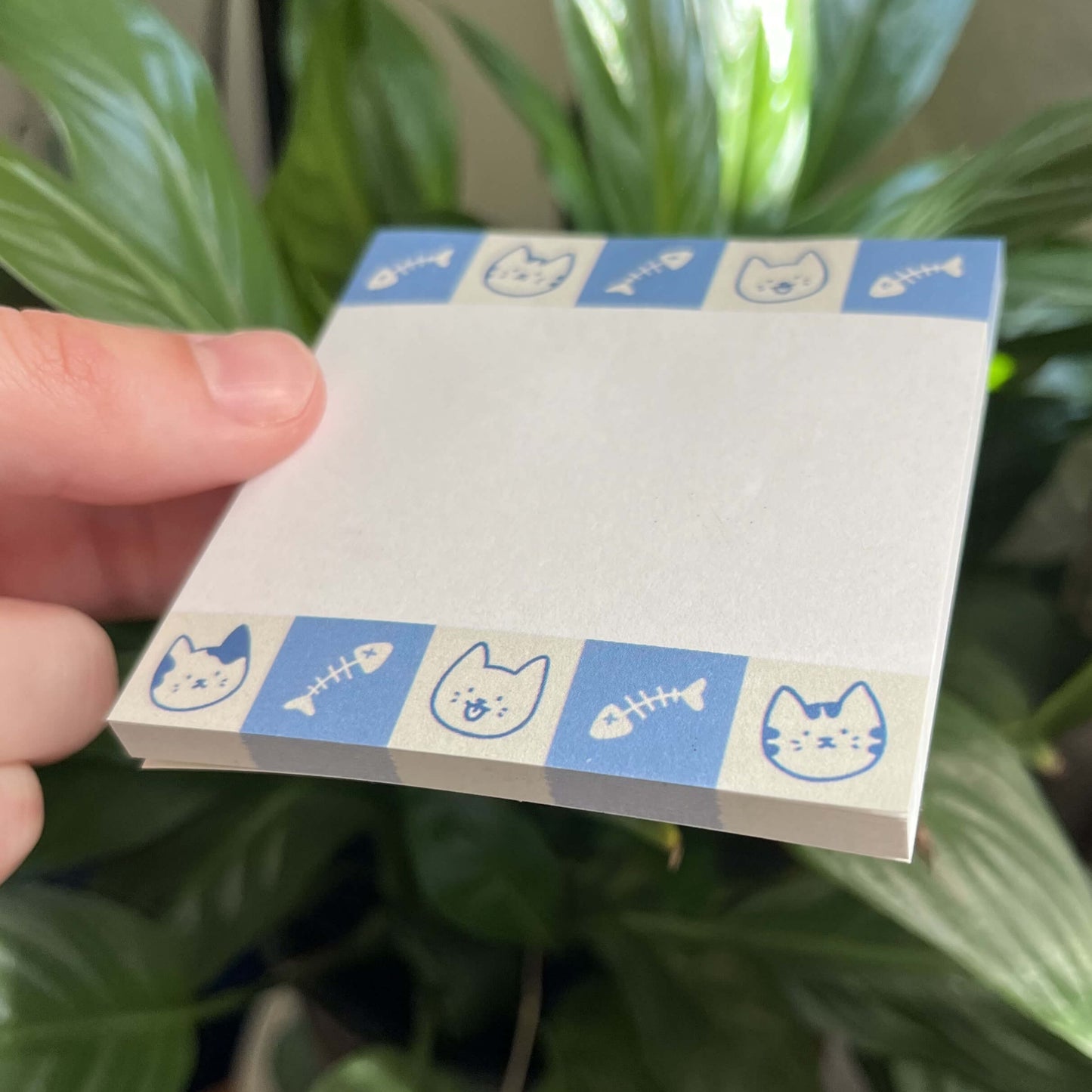 One square sticky notepad at an angle against a green back. The sticky note has blue borders featuring cat faces and fish bones.