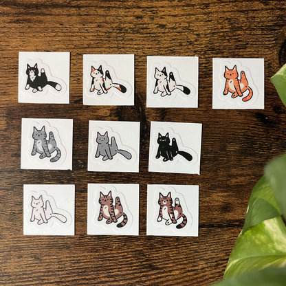 A set of 10 cat-themed stickers lined up against a wood background with leaves in the corner. Each cat is a different color and has its leg stuck up in the air, caught in the action of licking.
