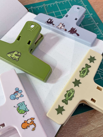 Four plastic clips in green, pale blue, cream, and white, each decorated with small animal or nature illustrations, arranged on a dotted notepad over a green cutting mat.
