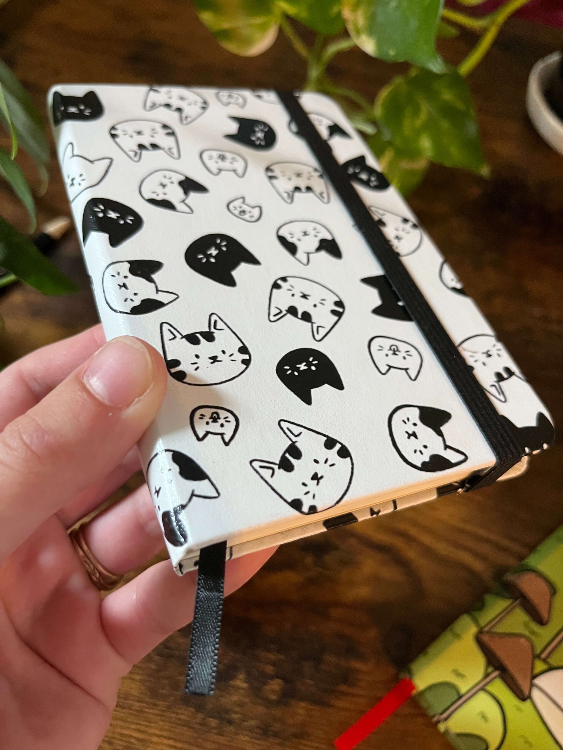 A notebook with a white cover featuring a pattern of various black and white cat faces, tied with a black elastic band and held over a wood table. 