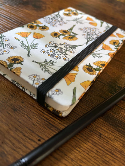 An elegant notebook at an angle with floral design featuring sunflowers, poppies, and daisies, lies on a rustic wooden table. The notebook is bound with a black elastic band.