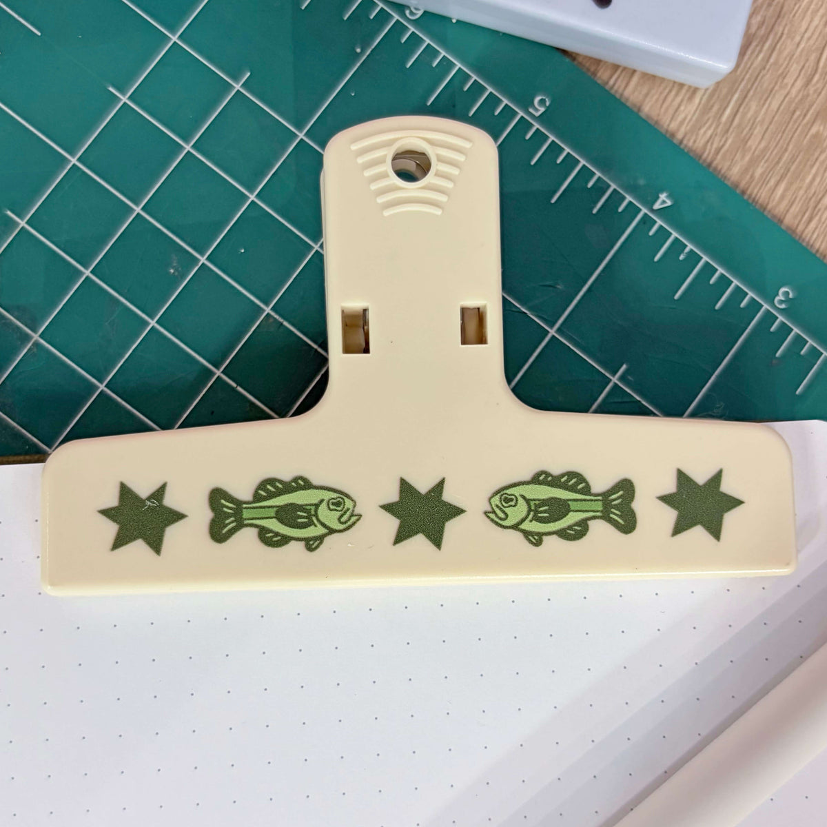 A cream-colored plastic binder clip decorated with green fish and star designs, resting on a green cutting mat over a white notebook.