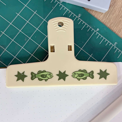 A cream-colored plastic binder clip decorated with green fish and star designs, resting on a green cutting mat over a white notebook.