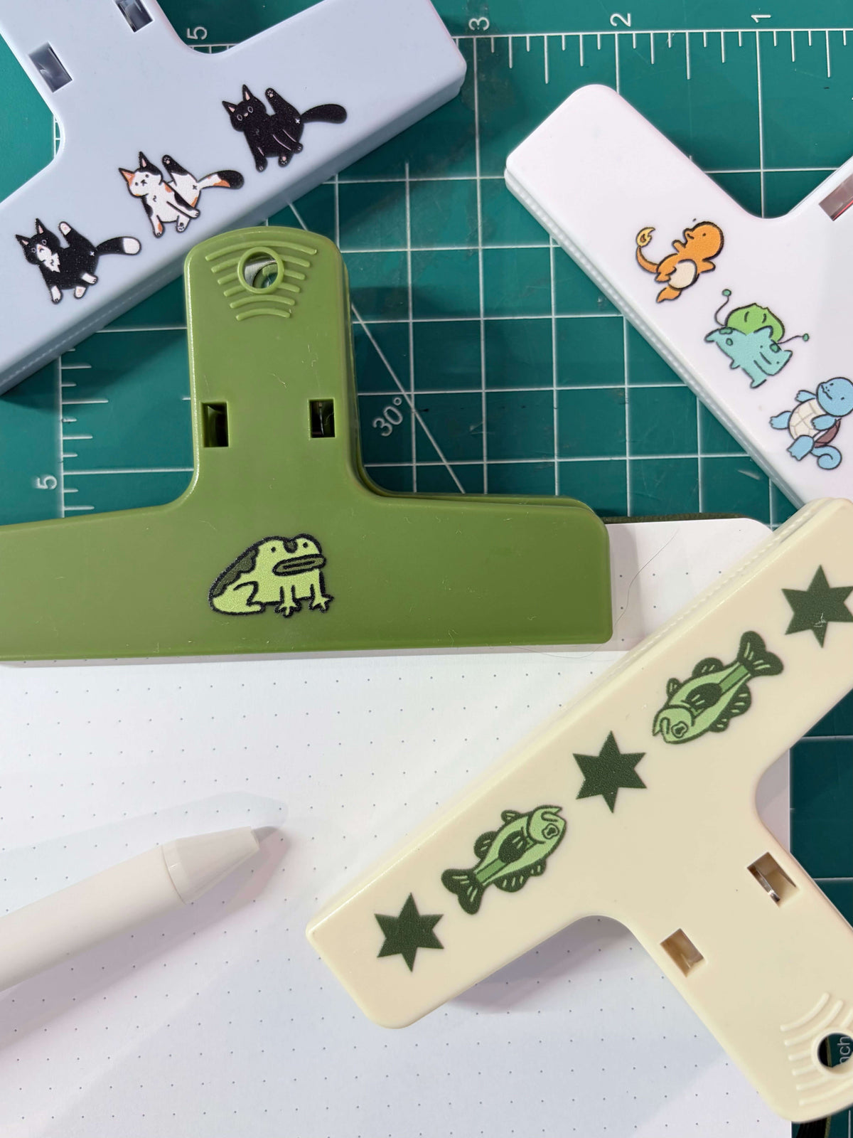 A set of T-shaped paper clips in different colors (green, cream, light blue) each decorated with a cute cartoon animal or nature motif (frog, fish, cat). The clips are arranged on a green cutting mat with a white dotted notebook and a white pen.