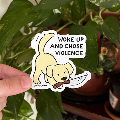 Hand holding a sticker with a dog and text 'Woke up and chose violence' in front of plants and books.