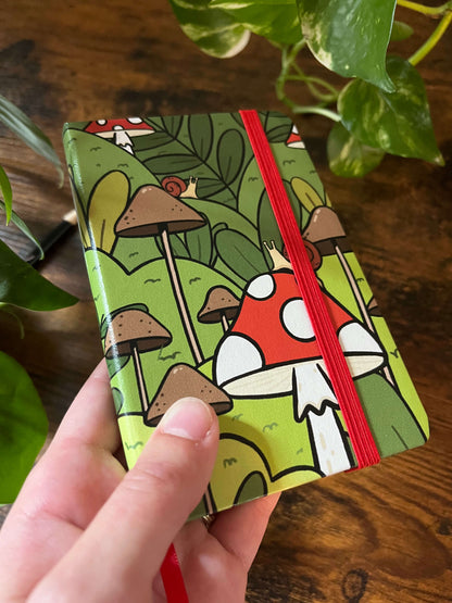 Angled notebook with red strap and cover of cartoon forest scene with red and white mushrooms, brown mushrooms, and lush green foliage, with two small snails sit on a mushroom.