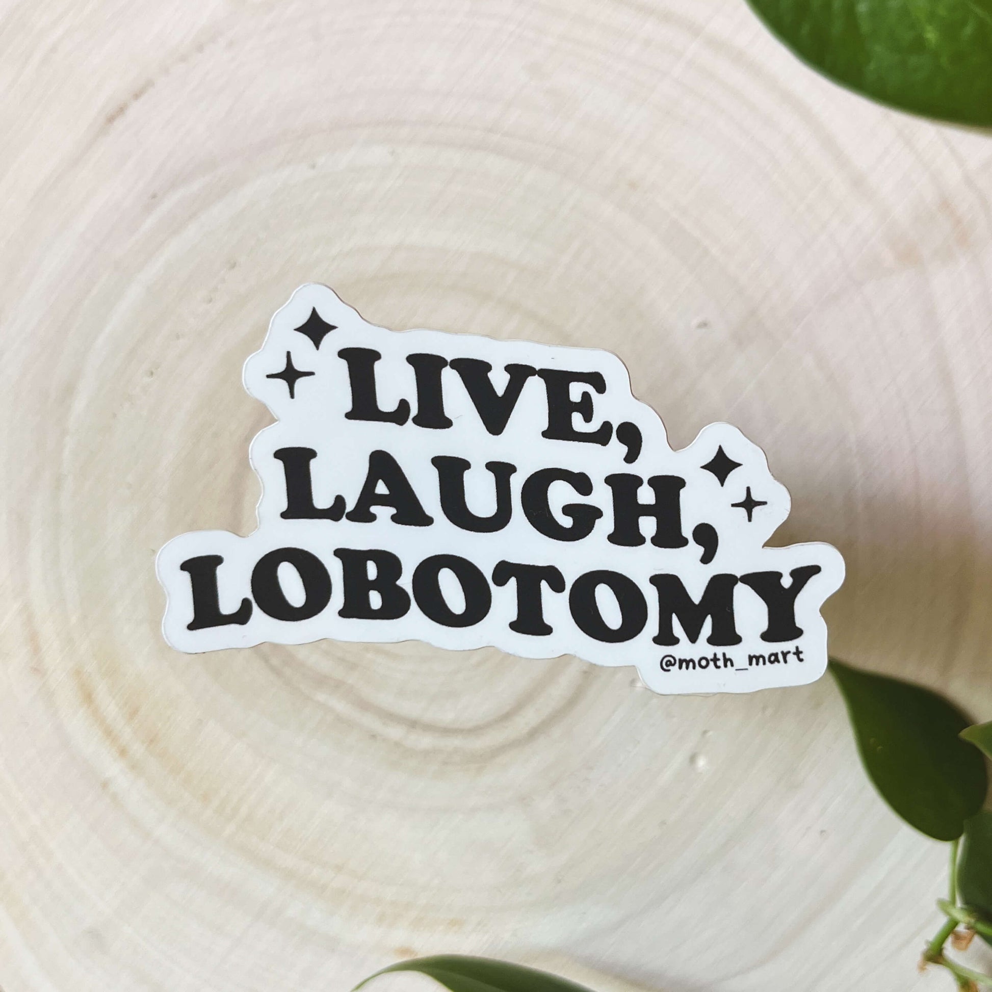 Sticker reading 'Live, Laugh, Lobotomy" on a wood background with green leaves