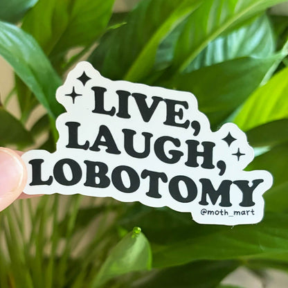 A die-cut sticker held by a person's finger against a background of green houseplant leaves. The sticker has a white background with bold black text reading 'LIVE, LAUGH, LOBOTOMY' and small star accents, plus the handle '@moth_mart' in the bottom right.