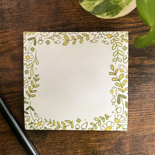 Square white sticky note pad with a cute floral border. Green and yellow leaves, flowers, and mushrooms decorate the edges, adding a cheerful touch.