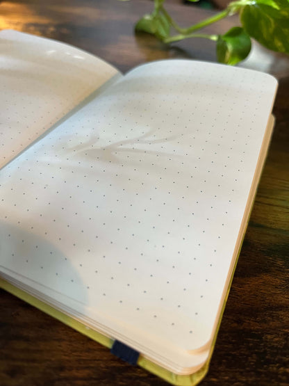 Open dotted notebook on a wooden table, sunlight casting soft leaf shadows on the pages.