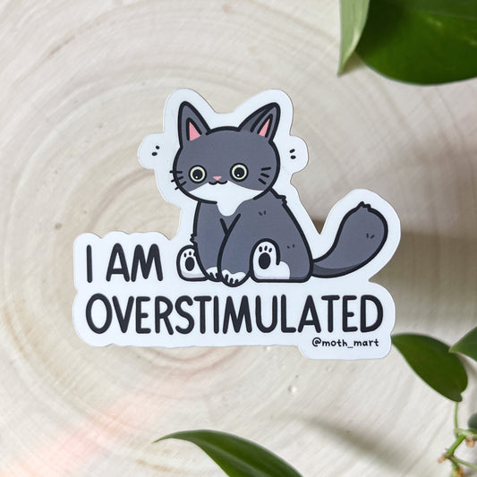Sticker with a cat illustration and text 'I AM OVERSTIMULATED' on a white background with green leaves.