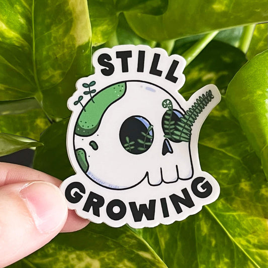 Sticker with the text "STILL GROWING" framed around a cartoon skull with ferns coming out of the eyes and moss growing on the back. 
