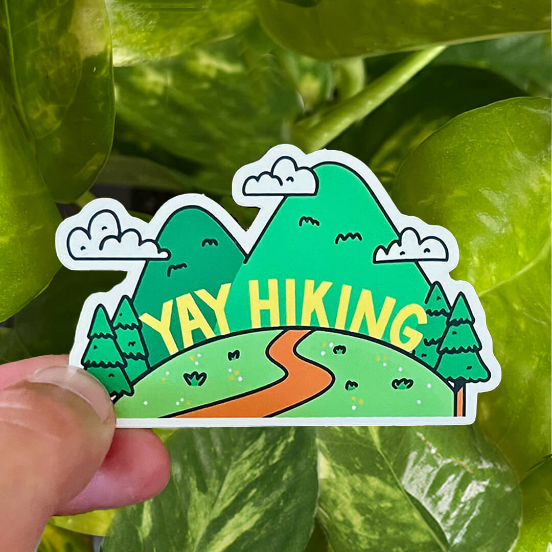 Sticker with text "YAY HIKING" on a green hill with hiking path, green trees and mountains.