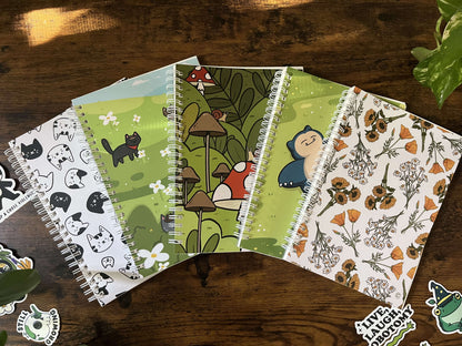 Five spiral bound reusable sticker books on a wood table. The covers feature black cats, a green field with flowers, mushrooms, a blue cartoon character, and floral patterns. Stickers are scattered around.
