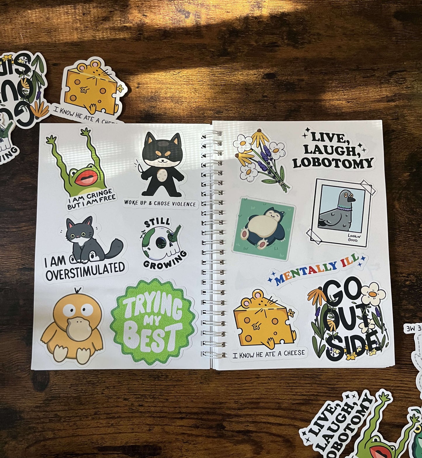 A sticker book open on a wood table. The left page displays a cat, a frog, and encouraging phrases. The right page features flowers, a pigeon, and playful messages. 
