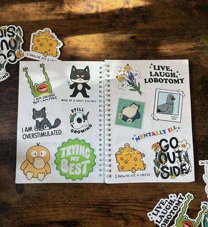 A sticker book open on a wood table. The left page displays a cat, a frog, and encouraging phrases. The right page features flowers, a pigeon, and playful messages. 
