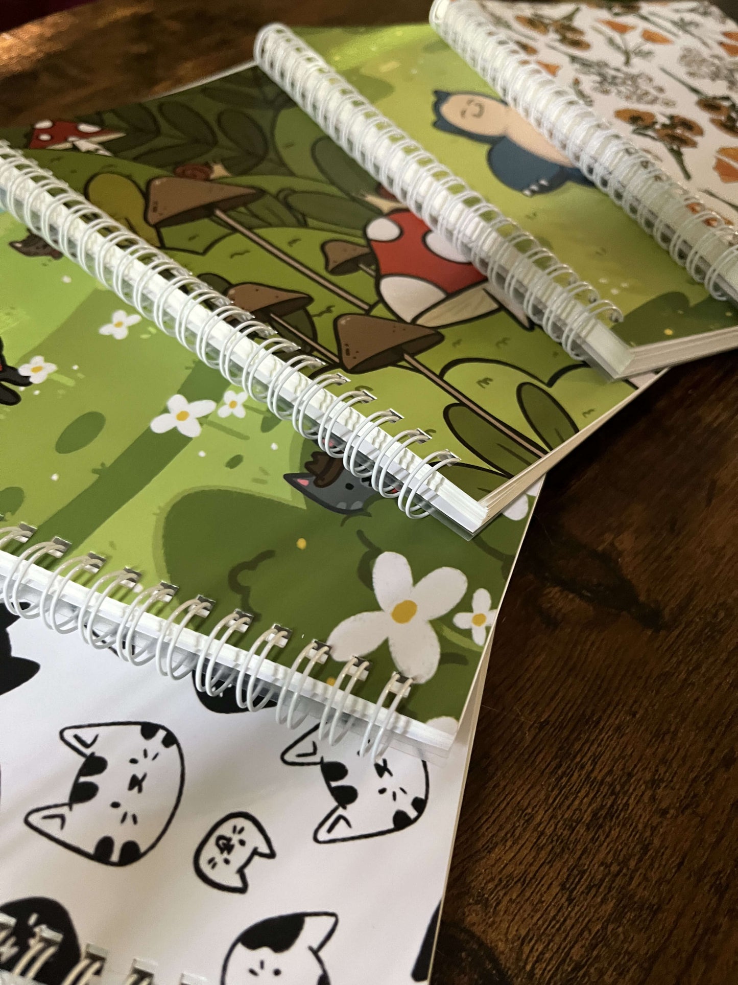 5 sticker books on a wood table at and angle. The covers feature black cats, a green field with flowers, mushrooms, a blue cartoon character, and floral patterns. Stickers are scattered around, creating a playful tone.