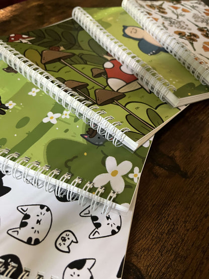 5 sticker books on a wood table at and angle. The covers feature black cats, a green field with flowers, mushrooms, a blue cartoon character, and floral patterns. Stickers are scattered around, creating a playful tone.