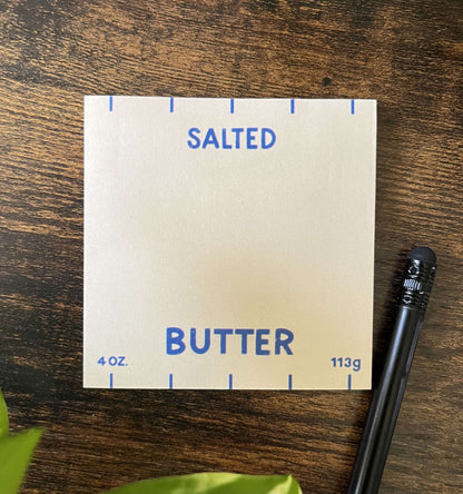 Butter-themed sticky notes on wooden table. The notes resemble a butter package, featuring blue text and markings that read "Salted Butter". A black pencil lies nearby.