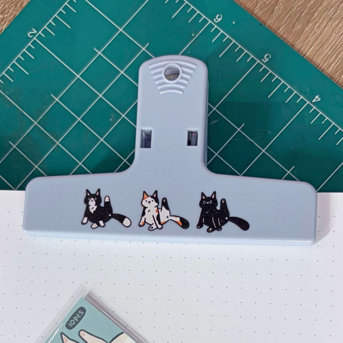 A light blue plastic binder clip decorated with three cute cartoon cat illustrations, resting on a green cutting mat over a white notebook.