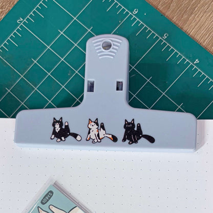 A light blue plastic binder clip decorated with three cute cartoon cat illustrations, resting on a green cutting mat over a white notebook.